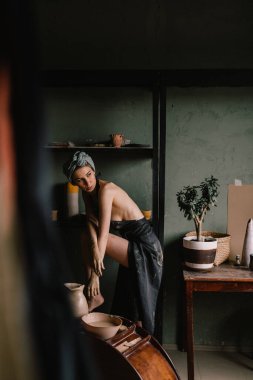 Kafasında bandaj ve çıplak vücudunda siyah bir önlükle güzel heykeltraş kız büyük pencerenin yanındaki çömlekçilik atölyesinde duruyor. konsept güzellik