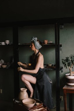 Kafasında bandaj ve çıplak vücudunda siyah bir önlükle güzel heykeltraş kız büyük pencerenin yanındaki çömlekçilik atölyesinde duruyor. konsept güzellik