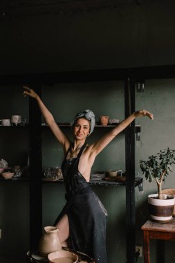 Kafasında bandaj ve çıplak vücudunda siyah bir önlükle güzel heykeltraş kız büyük pencerenin yanındaki çömlekçilik atölyesinde duruyor. konsept güzellik