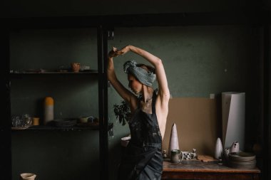 Kafasında bandaj ve çıplak vücudunda siyah bir önlükle güzel heykeltraş kız büyük pencerenin yanındaki çömlekçilik atölyesinde duruyor. konsept güzellik