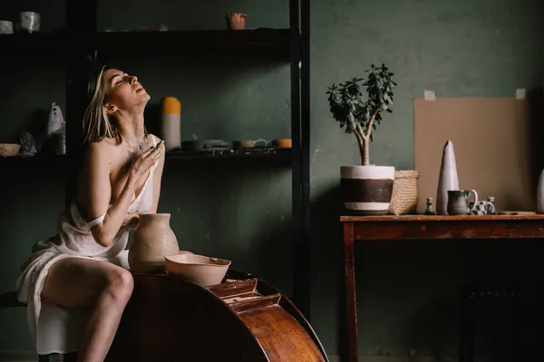 才华横溢的雕塑家女孩穿着薄薄的浅色睡衣 在靠近大窗户的陶瓷作坊里雕琢了一壶粘土 雕塑的概念 免版税图库照片