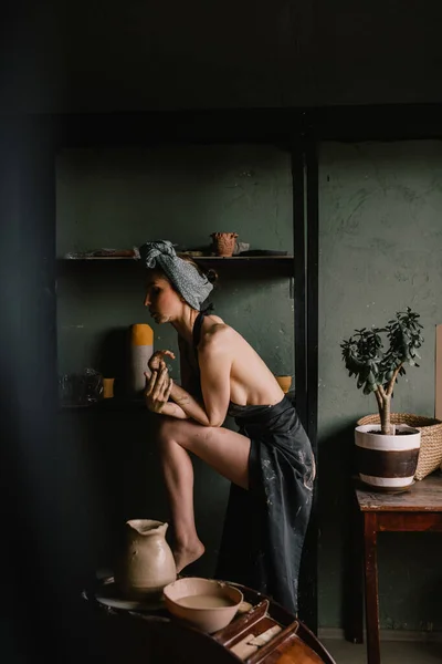 漂亮的雕塑家女孩头戴绷带 赤身裸体穿着黑色围裙 站在靠近大窗户的陶瓷作坊里 概念就是美 图库照片