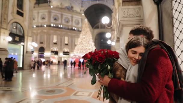 在圣诞树下的购物中心里 这位年轻人在求婚后拥抱了他的女朋友 恋爱中的一对浪漫的微笑的夫妇庆祝他们与花束的关系的某个日期 意大利米兰 — 图库视频影像