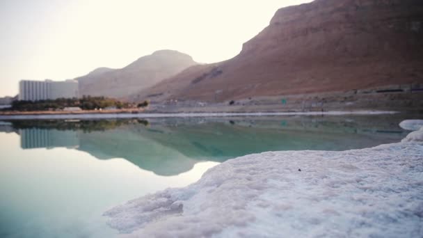 Arena completamente cubierta de sal cristalina parece hielo o nieve en la orilla del Mar Muerto, agua azul turquesa cerca, cielo coloreado con la distancia del sol de la mañana - paisaje típico en la playa de Ein Bokek, Israel — Vídeos de Stock