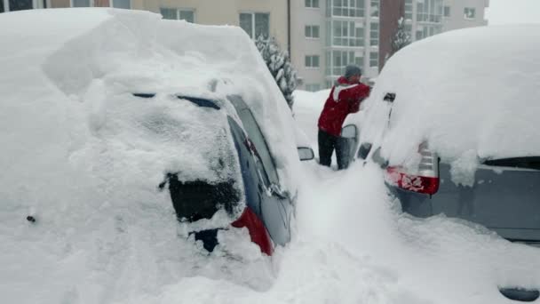 背景に雪の車を掃除している男がいる。雪のドリフトと嵐。駐車場に立っている車からブラシで雪をクリアバックパックにドライバ — ストック動画