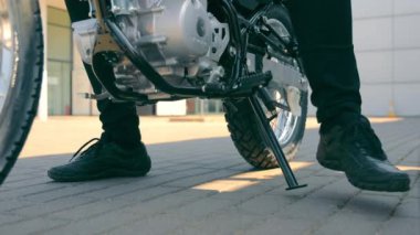 Motosikleti standa koyup motosikletten inen bir motosikletlinin bacakları. Motosikletli bir adam geldi ve otoparka koydu..