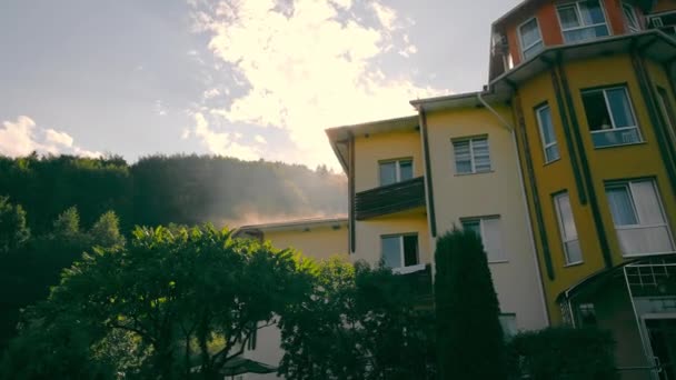 De zonnestralen van de ochtendzon die uit het huis komen. Zonsondergang na de regen. Het dak van het huis dampt na de regen. — Stockvideo