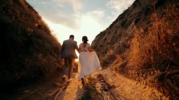 Um jovem casal caminha em direção ao sol entre duas montanhas. Pessoas felizes de mãos dadas e a caminhar em frente ao sol. Silhueta de um homem e uma mulher no balcão de sol. Tempo juntos. — Vídeo de Stock