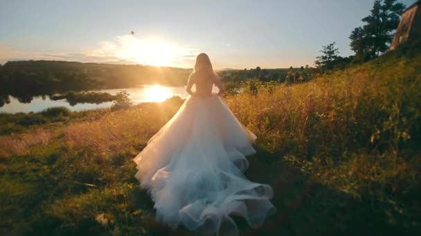 Красивая невеста бежит к озеру на фоне заката. Закат на Моранранранране и лучи на подсветке. молодая женщина в длинном шифонном платье — стоковое видео