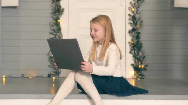 Een klein meisje dat een videogesprek voert met een laptop. Het meisje zit voor de deur van haar huis en communiceert via een videolink met een laptop op schoot. — Stockvideo