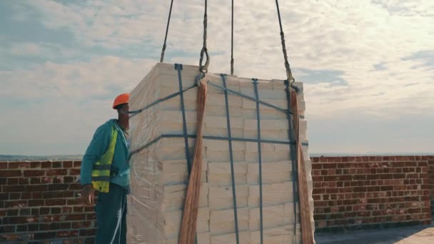 En byggkran lyfter en vit tegelsten och flyttar den runt byggarbetsplatsen. En byggarbetare hjälper till att transportera och lyfta en tegelkran. — Stockvideo