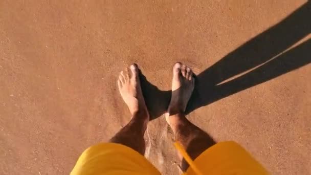 Pies masculinos de pie en la playa. Una ola que llega a una playa de arena lava los pies de un hombre. Vista superior de los pies de un hombre en una playa de arena — Vídeos de Stock