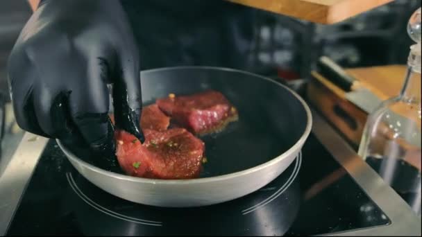 Schweinesteak in einer Pfanne kochen. Der Koch in schwarzen Handschuhen verteilt Schweinesteaks auf einer Pfanne aus nächster Nähe. — Stockvideo