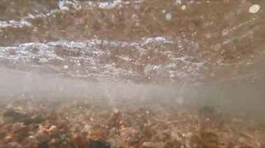 Suyun altındaki deniz dalgalarının arka planı. Dalgalar kıyı görüşüne yavaş çekimde geliyor. Suyun kıyıya akan gücü dipteki taşları kaldırır ve suda kabarcıklar oluşturur..