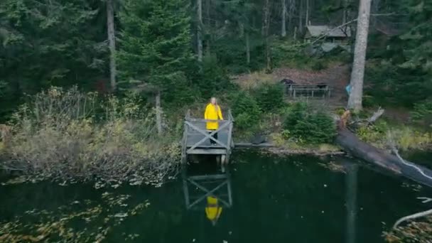 Una niña con un impermeable amarillo se encuentra junto al lago en un fabuloso bosque oscuro. Chica turística se encuentra en un puente cerca de un pequeño lago en las tierras altas. — Vídeos de Stock