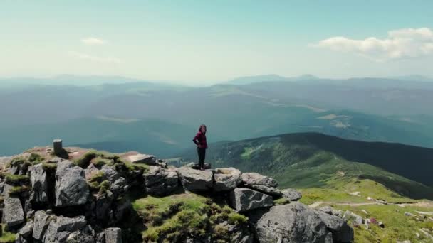 Una joven mujer se para en la cima de una montaña rocosa y levanta las manos, sonriendo. Paisaje escénico de montaña. Vista aérea. Mujer de pie en la cima de una montaña y mira a la distancia — Vídeo de stock