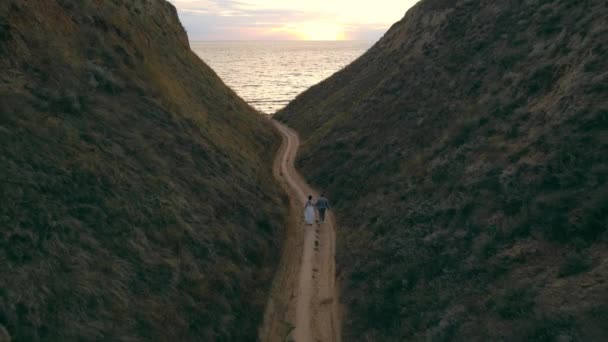 Vista desde el dron sobre un joven par de dos personas caminando hacia el mar al atardecer. Niño y niña caminan en las montañas junto al mar. — Vídeos de Stock