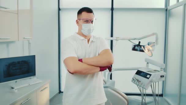 Un dentista es un hombre de nacionalidad caucásica. Un médico con máscara y gafas se para en el consultorio de dentistas de la clínica dental y mira a la cámara. — Vídeos de Stock
