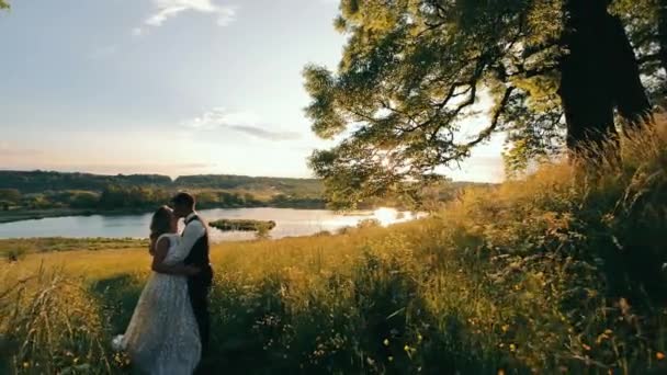Egy boldog fiatal pár egy órát tölt a tónál napnyugtakor. A nap meleg naplementéje és a tó természetének széle. A nő a karjában becsukta a szemét, és élvezi a fogadó pillanatok — Stock videók