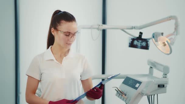 Fehér női fogorvos a fogorvosi rendelőben, átlátszó védőmaszkban és mappában a kezében. Lány fogorvos néz kamera. — Stock videók