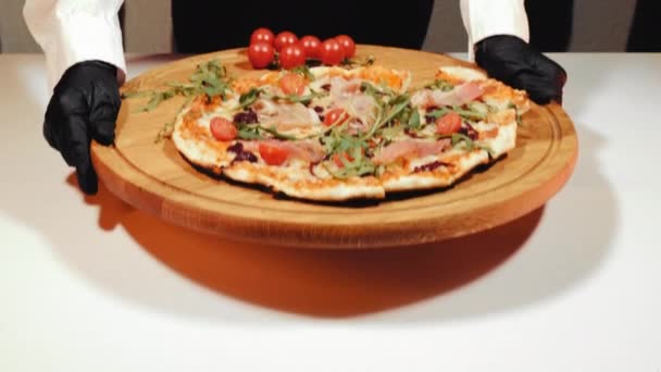 Le plat fini est une pizza maison avec des tomates, du prosciutto et des feuilles de roquette vertes que le serveur met sur la table blanche. Servir une pizza prête à l'emploi dans un restaurant. — Video