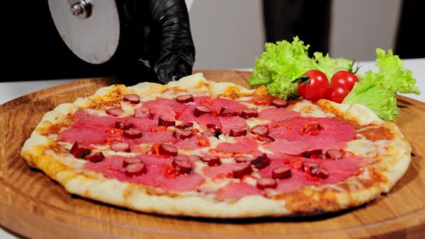 Cortar la pizza terminada con un cuchillo redondo. El camarero de cerca corta una rebanada de pizza con salchichas de caza. — Vídeo de stock