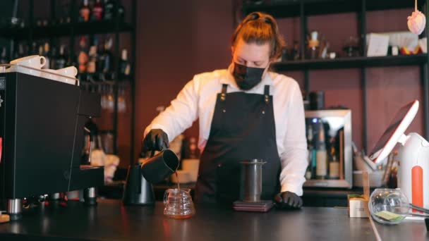 Koruyucu maskeli kadın barista kahve hazırlıyor, kafede bardağa sıcak su dolduruyor. At kuyruklu esmer kız puf yapıyor, elinde kahve demliği var. Çalışma yeri kavramı — Stok video