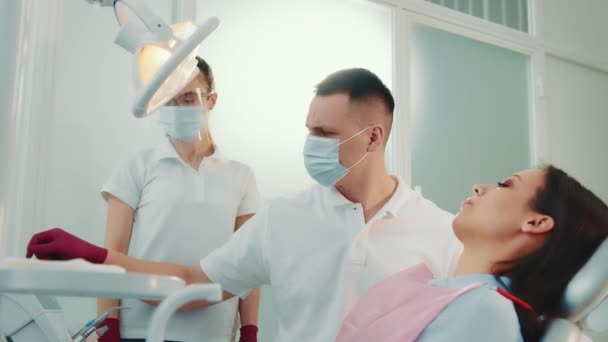 Vista lateral del dentista y asistente en el tratamiento uniforme de dientes de paciente femenina con espejo que trabaja en clínica dental. Doctor sosteniendo espejo dental, curando dientes de mujer bonita. Concepto de odontología — Vídeos de Stock