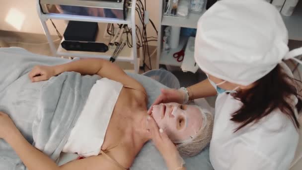 Sidovy av kosmetolog i mask gör nackmassage till vuxen kvinna patient i skönhetssalong. Ovanför utsikt över kaukasisk kvinna får ansikte och hals massage, avkopplande på soffan. Begreppet skönhetsvård — Stockvideo