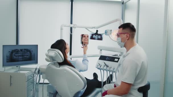 Молодая женщина сидит на стуле, смотрит в зеркало, улыбается после стоматологической процедуры. Врач и пациент исследуют зубы с зеркалом после лечения в стоматологической клинике. Концепция стоматологии — стоковое видео