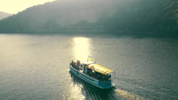 Passenger ferry slowly sailng on calm river surface along hilly banks in sunny day. Aerial view of ferry boat with blue and yelloow flag on top, floating on wide, wavy river. Concept of recreation — Stock Video