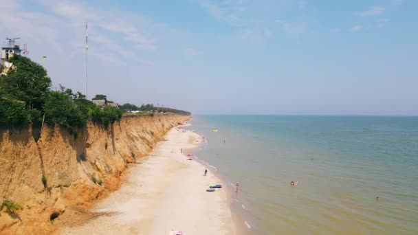 Flying over sandy beach and blue transparent water of sea, along waterfront in summer. Aerial view from drone of steep seashore, beach with chairs, sun umbrellas and people swimming. Concept of relax — Stock Video