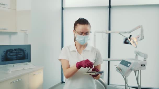 Zahnärztin mit Schutzmaske, die in der Klinik einen intraoralen Spiegel aus dem Tablett nimmt. Porträtansicht eines hübschen Assistenten in Handschuhen, der Zahnwerkzeug auswählt und die Kamera im Innenraum betrachtet. Konzept der Zahnmedizin — Stockvideo