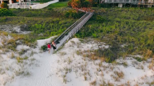 Zwei Mädchen gehen in der Abenddämmerung auf einem Holzsteg vom Sandstrand weg. Luftaufnahme von Mutter und Tochter, die zu Fuß über eine Fußgängerbrücke gehen und nach Hause zurückkehren, nachdem sie sich am Strand ausgeruht haben. Konzept der Entspannung — Stockvideo