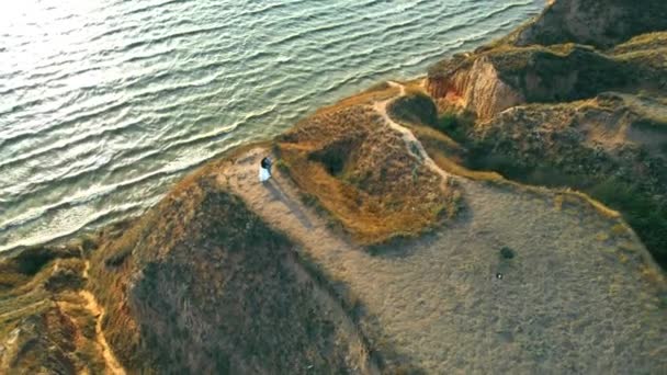 Ein junges Paar, das dicht an dicht auf einem hohen Hügel am Meer steht und sich in der Sonne umarmt. Luftaufnahme von der Drohne eines verliebten Mannes und einer verliebten Frau, die auf einer Klippe über der Meeresoberfläche im Licht des Sonnenuntergangs stehen. Konzept der Romantik — Stockvideo