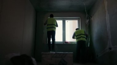 Alçı boyacılarının siluetleri odanın penceresinin yakınına sıva yapıştırıyor. Karanlık oda ve odada tamirat yapan iki işçi silueti.