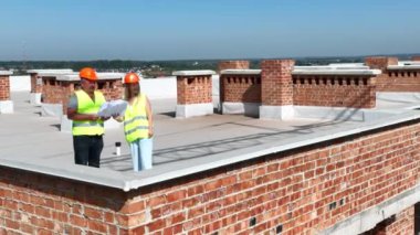 Hava manzaralı iki mimar, bir adam ve bir kadın, çok katlı tuğla bir binanın çatısındaki inşaat planını inceliyorlar.