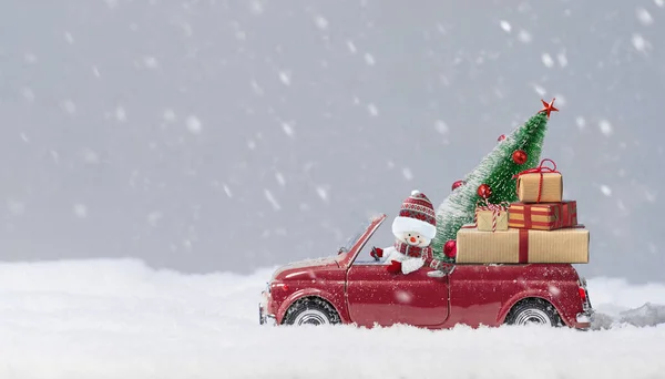 Schneemann in rotem Auto liefert Weihnachtsbaum und Geschenke vor verschneitem Hintergrund. — Stockfoto