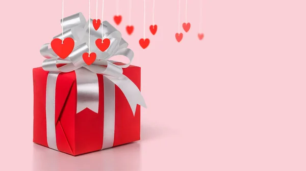 Red Gift box with silver bow and red hearts on a ropes on a pink background. — Fotografia de Stock