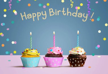 Happy Birthday card. Cupcakes with burning candles on the pink table and blue wall background with confetti