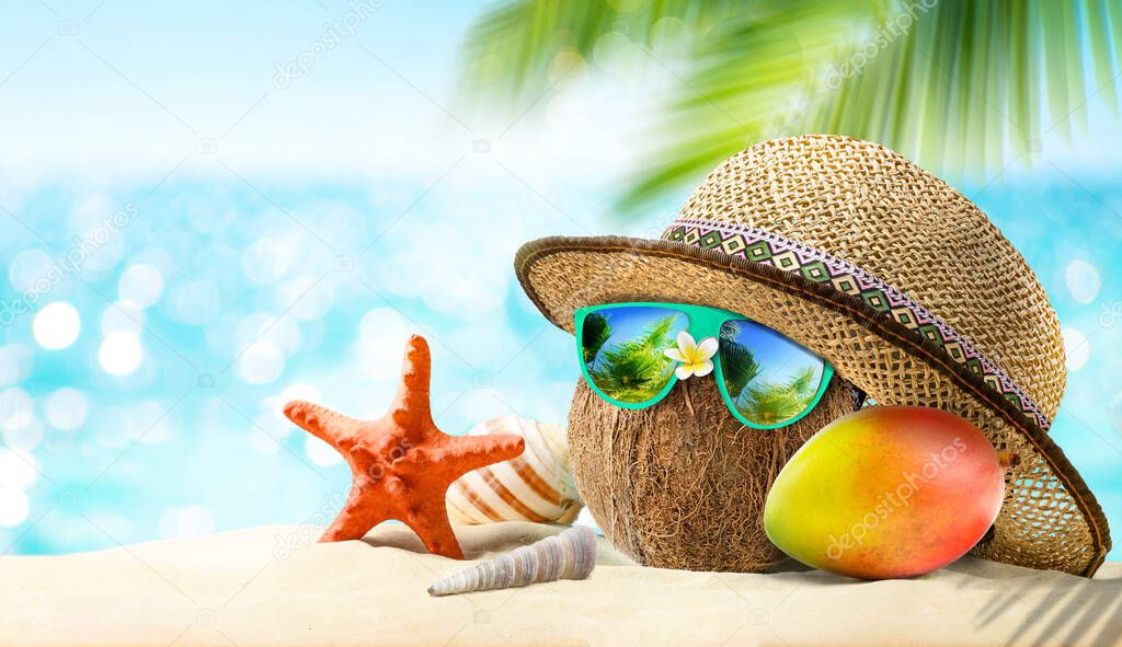 Summer Vacation Concept. Coconut with sunglasses and straw hat at tropical beach with starfish, mango fruit and sea background.