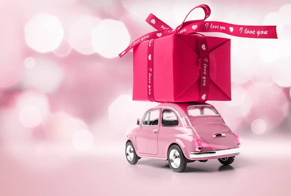 Pink retro toy car delivering red gift box with ribbon on pink background with nice bokeh. February 14 card, Valentine\'s day. 8 March, International Happy Women\'s Day. I love you text
