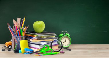 Okul malzemeleri arasında çalar saat ve ahşap masa üzerinde elma ve yeşil karatahta arka plan var. Boşluğu kopyala