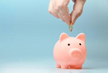 Hand putting coin to pink piggy bank on a blue background. Copy space