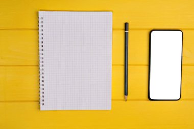 Top view of a notepad with a pencil and a blank smartphone on a yellow wooden background.White empty space for advertise text