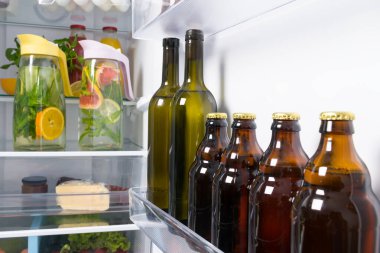 Beyaz bir buzdolabının arka planında, şarap şişeleri, bira ve cam sürahiler narenciye meyveleri raflarda yatıyor.