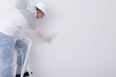 Koruyucu giysi ve kask giyen bir müteahhit, duvarlardaki fazla yapıştırıcıyı çıkarmak için duvar kağıdını plastik spatulayla aynı seviyeye getiriyor.