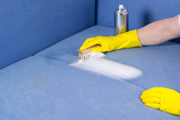 hands in yellow gloves, apply a cleaning agent from a metal balloon to the blue fabric of the sofa
