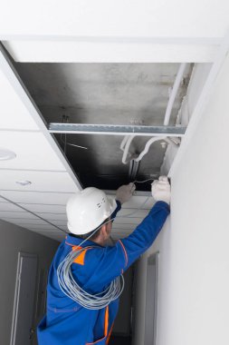 Mavi üniformalı ve beyaz kasklı bir usta, kablosuz ağ yönlendiricisinin kablolarını sahte tavan sistemine yerleştiriyor.
