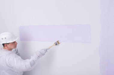 Koruyucu giysili ve kasklı bir ressam, açık mor renkli beyaz bir duvar boyuyor, yazacak bir yer var, yan görüş var.